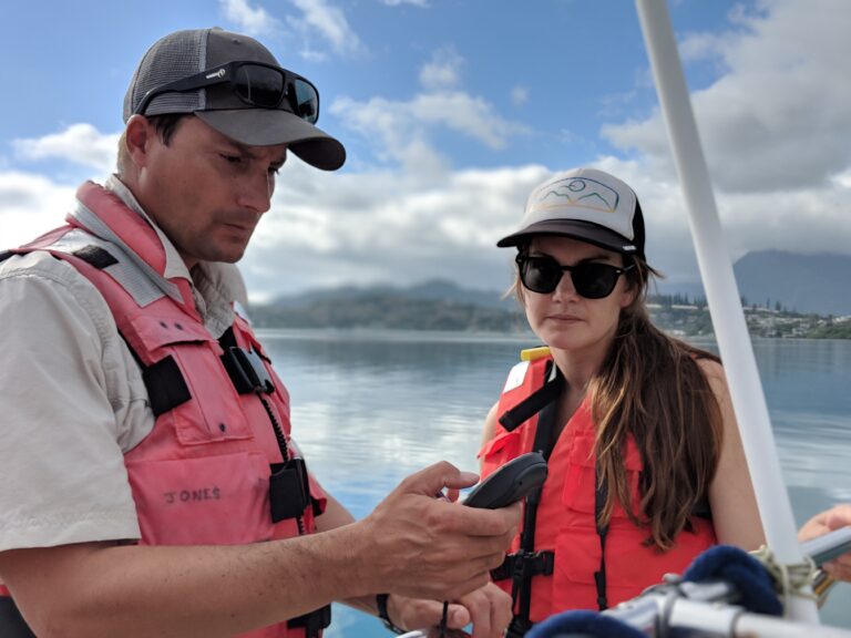 Two people in life vests on a boat, one holding a device, with water and mountains in the background.