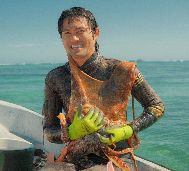 A smiling diver in wetsuit and gloves holds a large octopus on a boat with the ocean in the background.