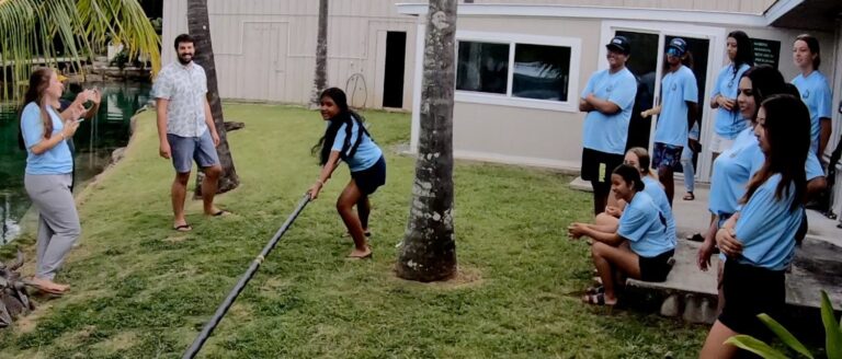 A group of people in blue shirts watch a woman using a long pole near a lawn and building.