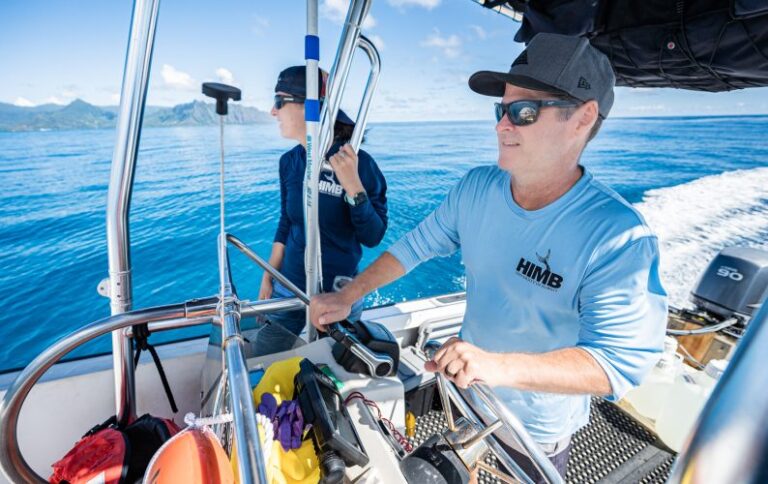 Two people in sunglasses driving a small boat on clear blue water, with mountains in the background.