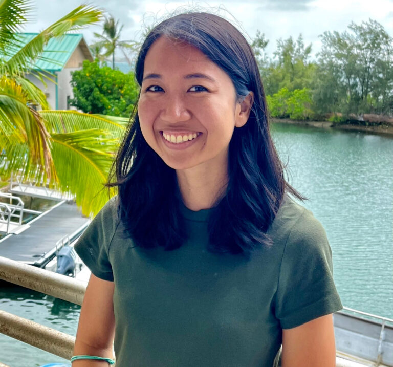 Woman smiling outdoors by water, with boats, palm trees, and greenery in the background.