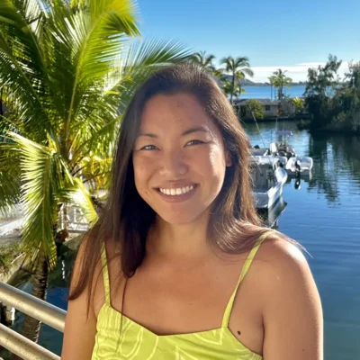 Woman smiling outdoors near palm trees, water, and boats on a sunny day.