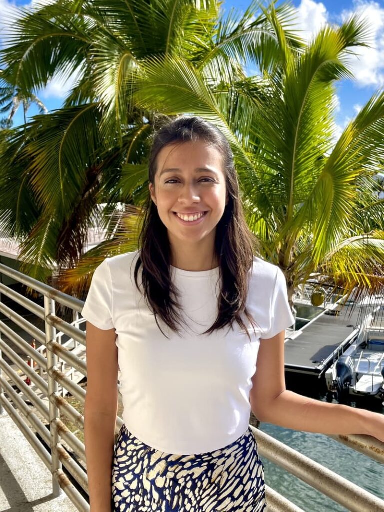 Woman smiling outdoors by palm trees and water, wearing a white shirt and patterned skirt on a sunny day.