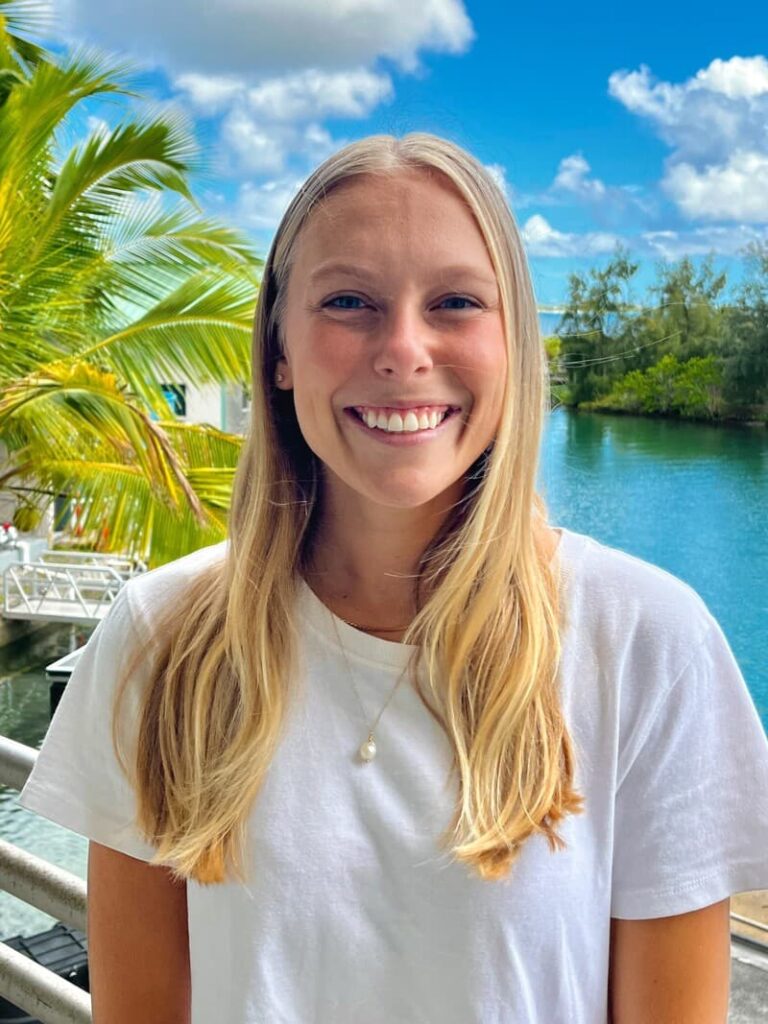 Smiling woman with long blonde hair in a white shirt stands by water and palm trees on a sunny day.