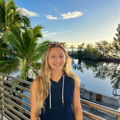 Smiling woman with long blonde hair stands by a waterfront railing with palm trees and blue sky in the background.