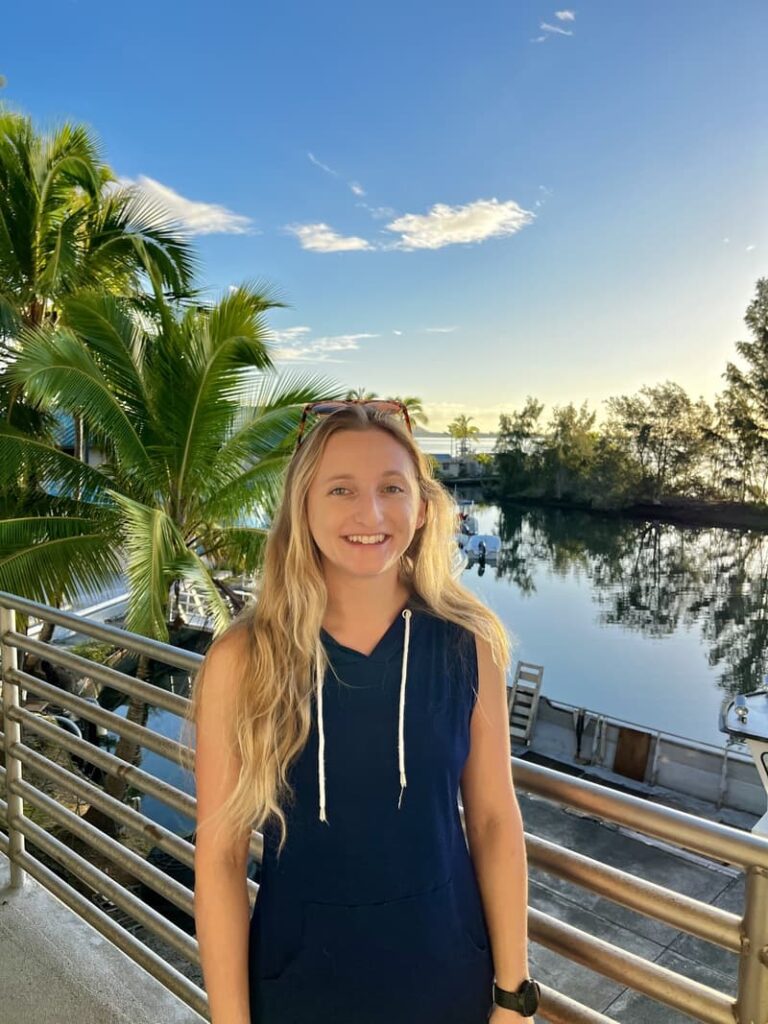 Smiling woman with long blonde hair stands by a waterfront railing with palm trees and blue sky in the background.