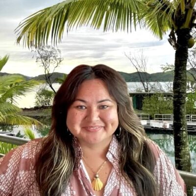 Woman smiling outdoors near palm trees and water, with a dock and greenery in the background.