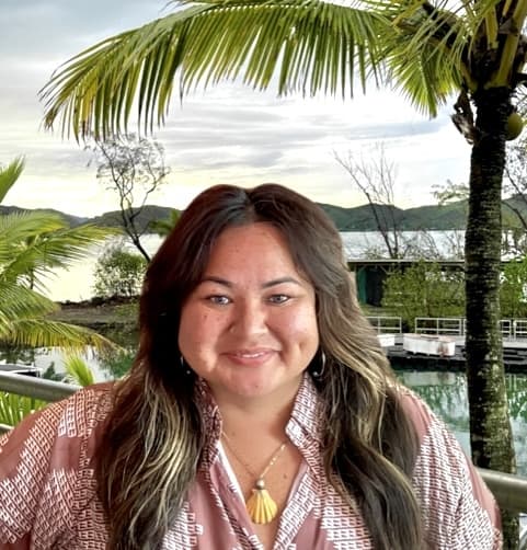 Woman smiling outdoors near palm trees and water, with a dock and greenery in the background.