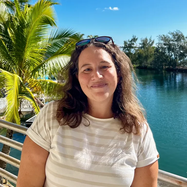 Woman smiling by a waterfront, with palm trees, blue sky, and sunglasses resting on her head.