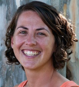 Woman with short, curly brown hair smiling in front of a tree with a mottled bark background.