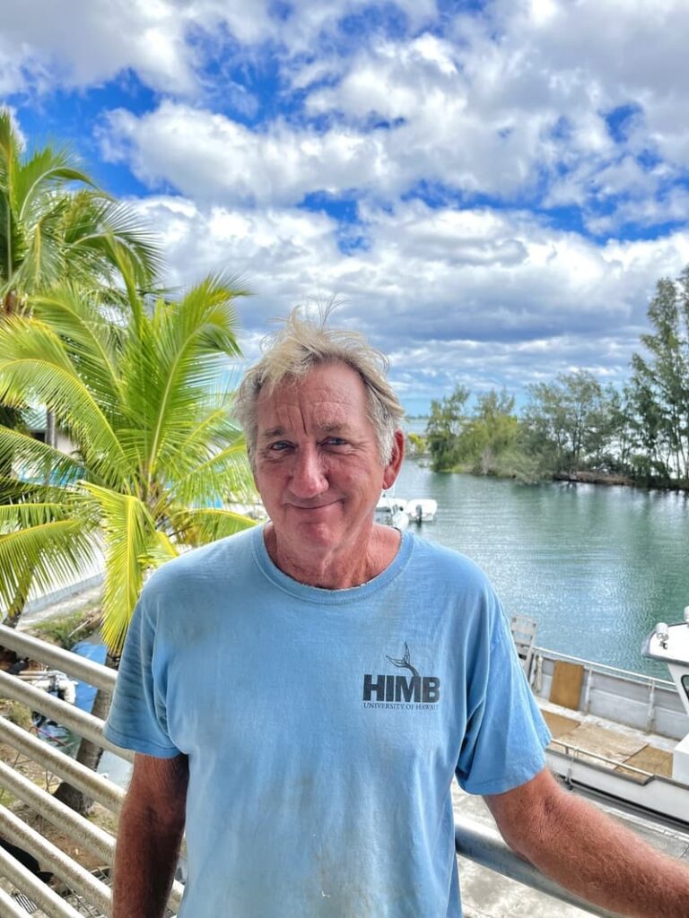Man in a blue HIMB shirt stands by water with boats, palm trees, and cloudy sky in the background.