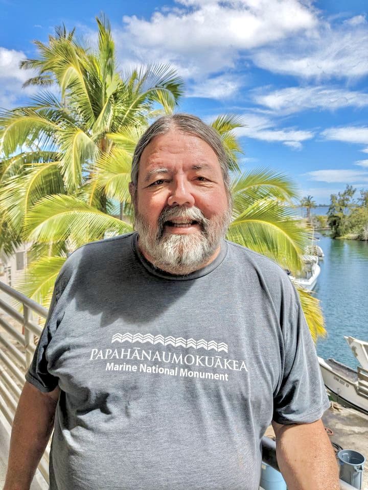 Smiling man with gray beard stands by palm trees and water, wearing a Papahānaumokuākea T-shirt.