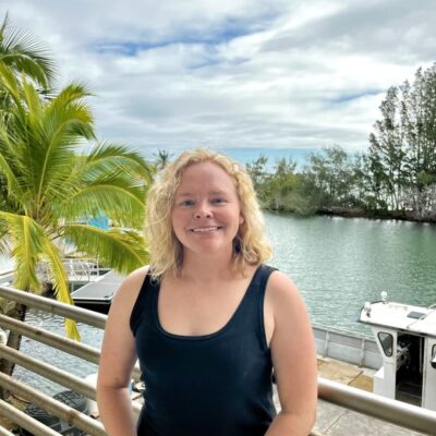 Woman smiling on a balcony by the water, with palm trees, boats, and cloudy sky in the background.