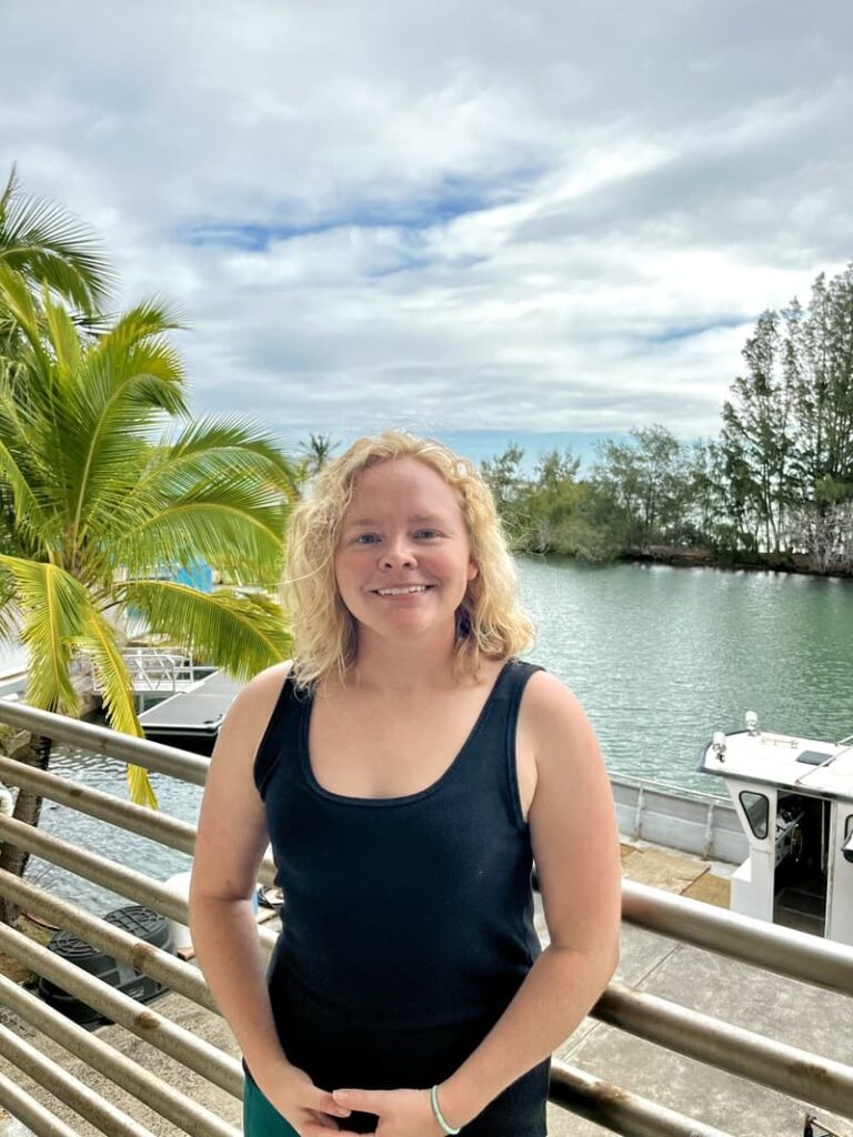 Woman smiling on a balcony by the water, with palm trees, boats, and cloudy sky in the background.