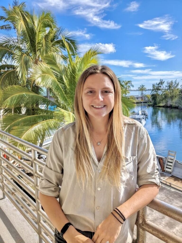 Smiling woman stands by a railing with palm trees and a marina in the sunny background.