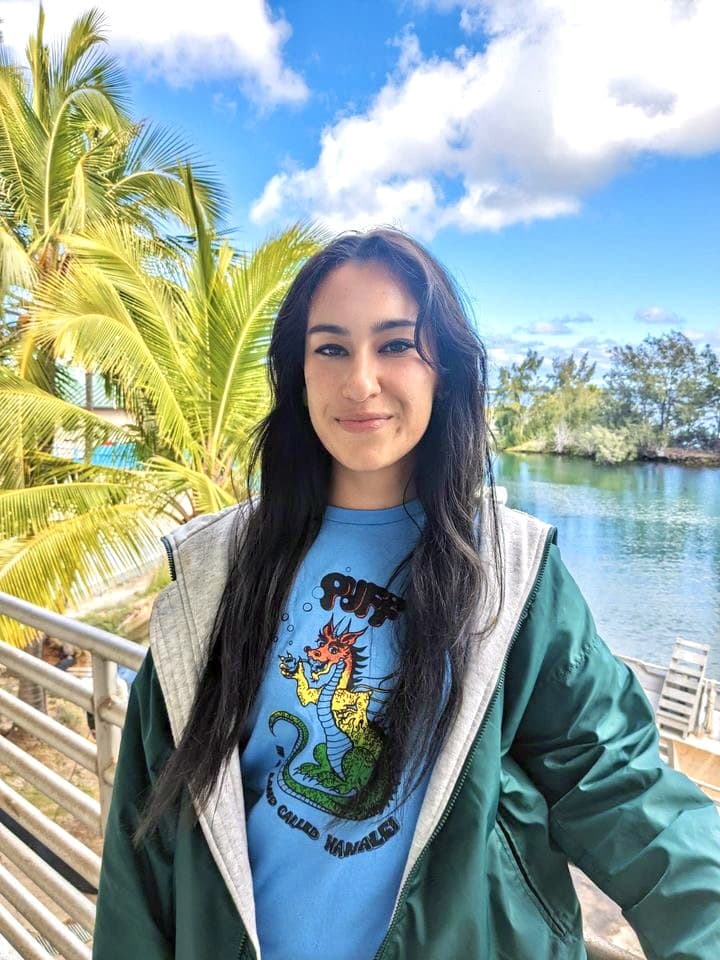 Woman with long dark hair smiles outdoors by water, palm trees, and blue sky, wearing a green jacket and dragon shirt.