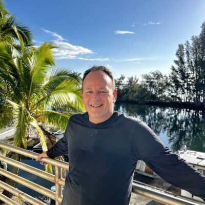 Smiling man in a black shirt stands by a railing with a palm tree and water in the background.