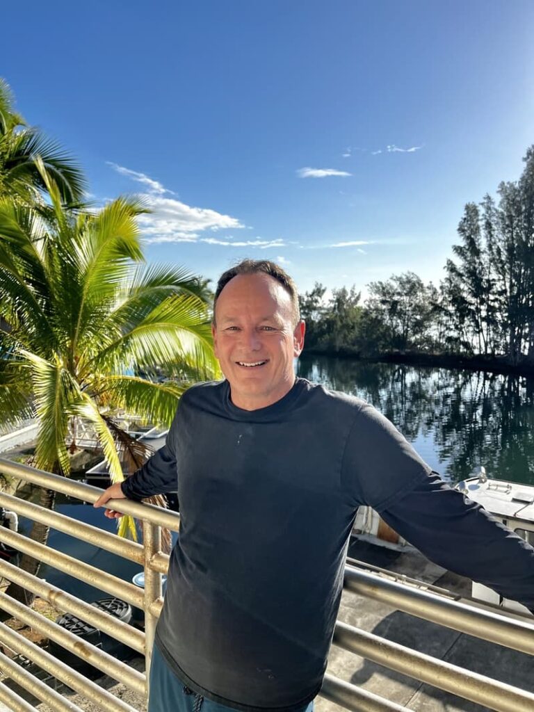 Smiling man in a black shirt stands by a railing with a palm tree and water in the background.