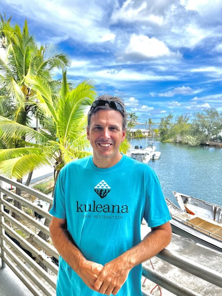 Smiling man in a blue "Kuleana Coral Restoration" shirt stands by a waterfront, with palm trees and boats behind him.