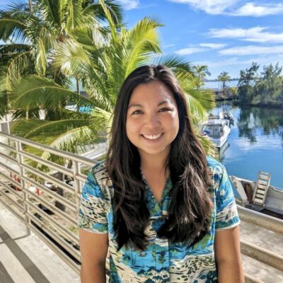 Woman smiling in a tropical shirt, standing by water with boats and palm trees on a sunny day.