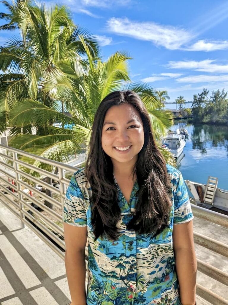 Woman smiling in a tropical shirt, standing by water with boats and palm trees on a sunny day.
