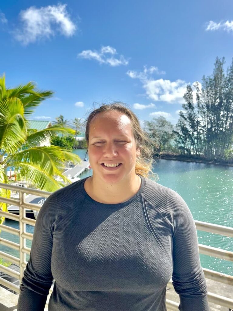 Smiling person in a gray shirt stands by water, palm trees, and blue sky with clouds in the background.