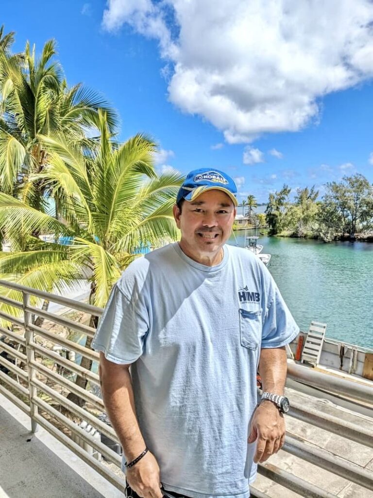 Man in a light blue shirt and blue cap smiling on a balcony by palm trees and water under a sunny sky.