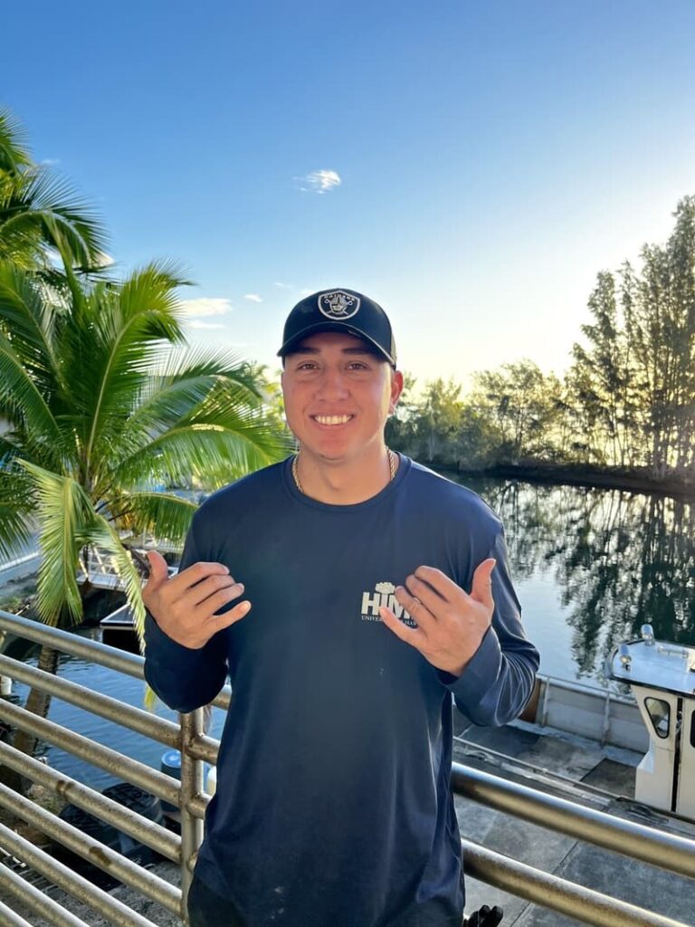 Smiling man in a dark shirt and cap stands by a railing, with water, trees, and blue sky in the background.