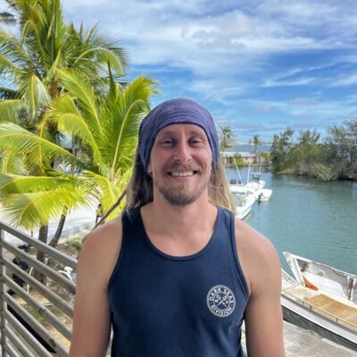 Smiling man in a tank top and headband standing by water, boats, and palm trees under a blue sky.