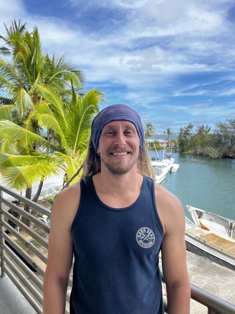 Smiling man in a tank top and headband standing by water, boats, and palm trees under a blue sky.