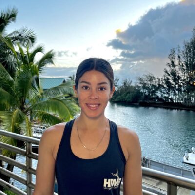 Woman in a black tank top smiles on a balcony by water with palm trees and a cloudy sky in the background.