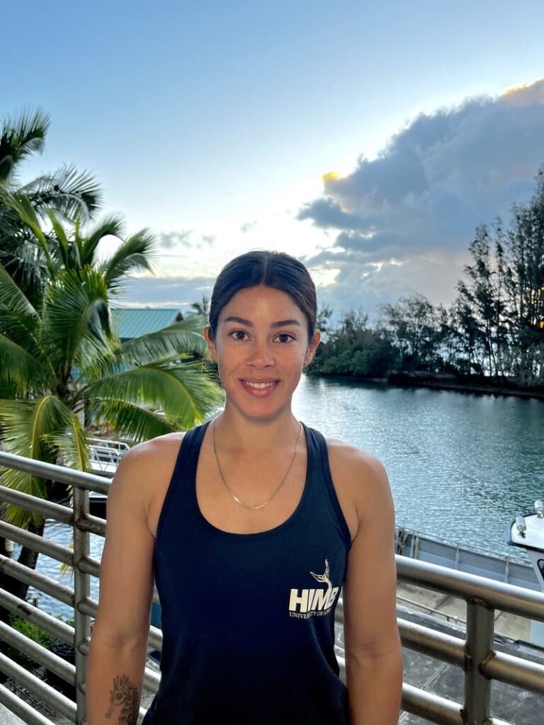 Woman in a black tank top smiles on a balcony by water with palm trees and a cloudy sky in the background.