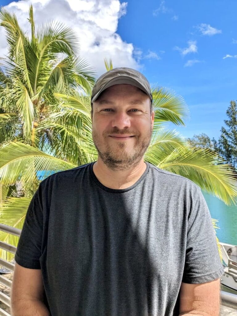 Smiling man in a gray shirt and hat stands outside with palm trees and blue sky in the background.