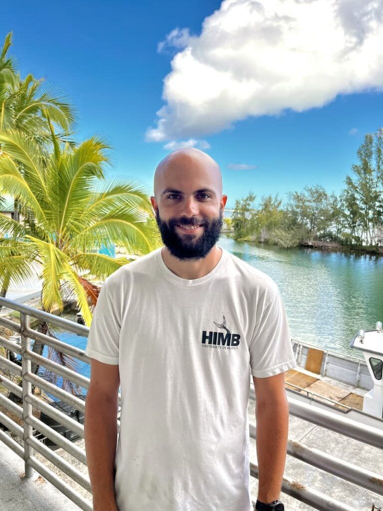 A man with a beard in a white HIMS t-shirt stands by water, palm trees, and a blue sky with clouds.