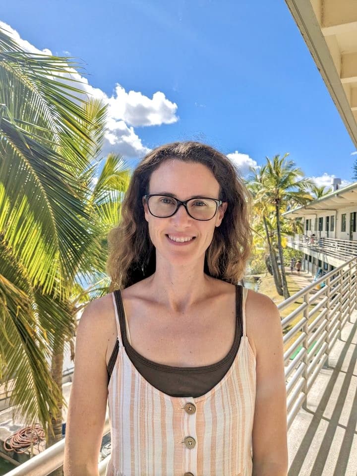 Woman with glasses smiling outdoors, standing by palm trees and a building under a bright blue sky.