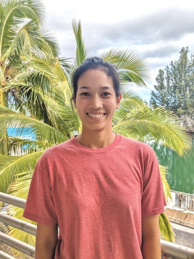 Smiling person in a red shirt stands outdoors with palm trees and water in the background.