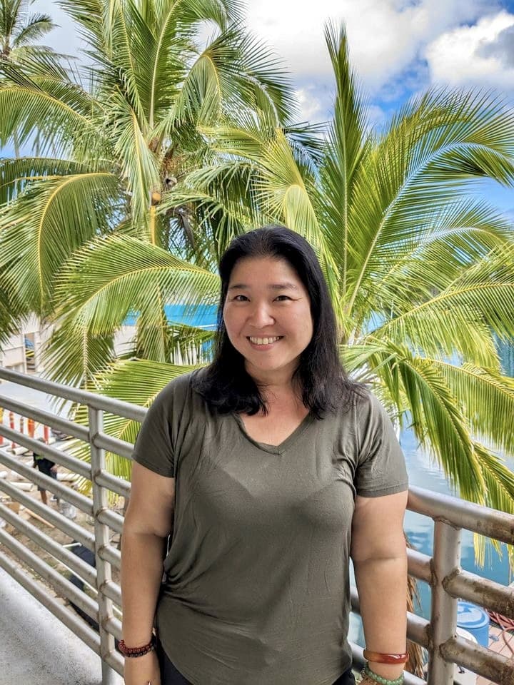 Smiling woman stands in front of palm trees and a sunny, blue sky near the ocean.