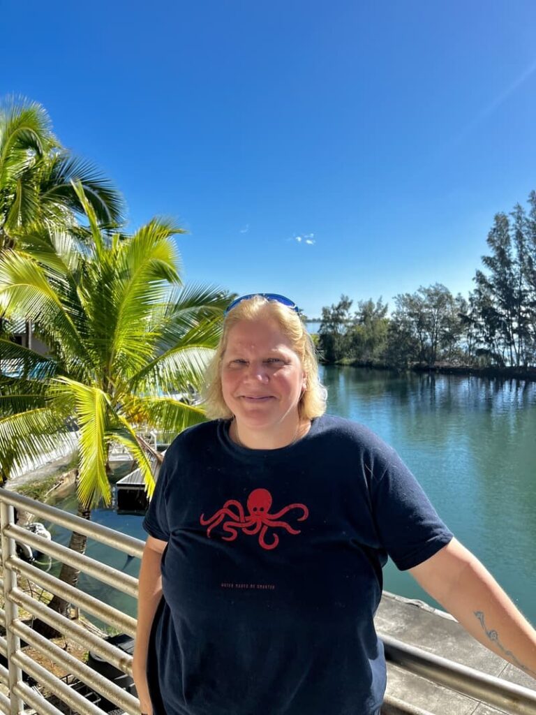 Woman standing by a waterfront railing, palm tree and blue sky in the background, wearing a black t-shirt.