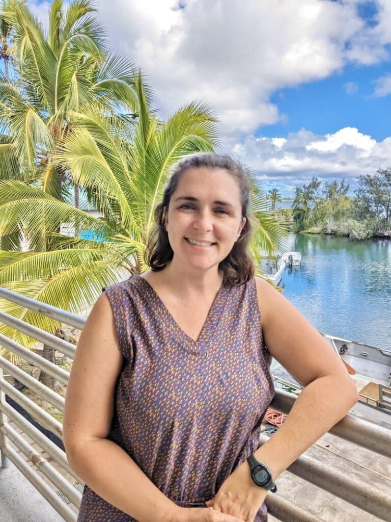 Woman smiling on a balcony with palm trees and water in the background under a partly cloudy sky.
