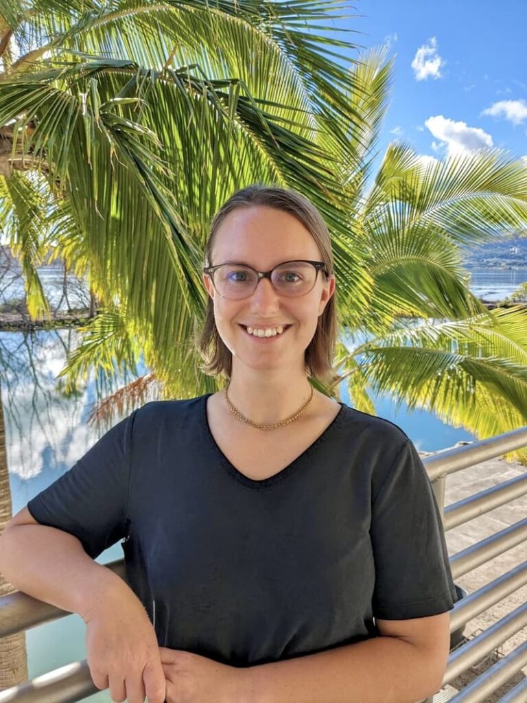 Smiling person with glasses stands in front of palm trees and water, wearing a black shirt.