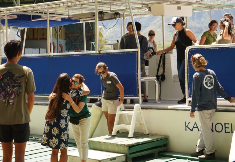 People greet each other and board a boat named Kaneohe, with mountains visible in the background.