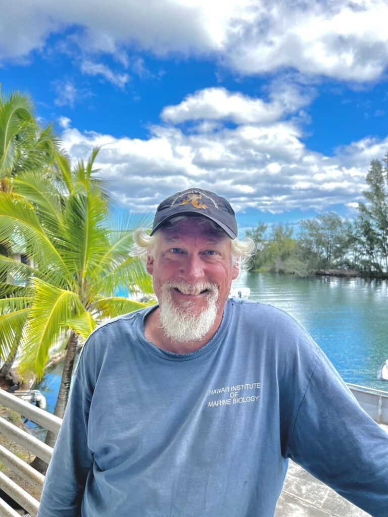 Smiling man with a white beard stands by water, palm trees, and blue sky, wearing a cap and blue shirt.