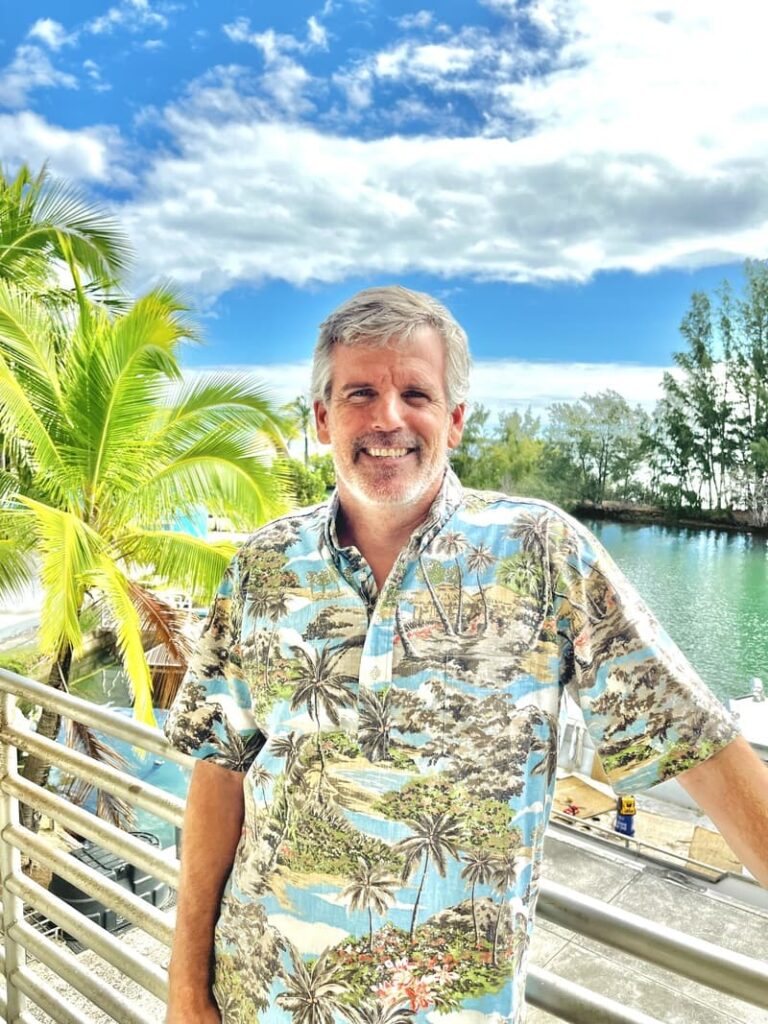 Smiling man in a tropical shirt stands by a railing with palm trees, water, and blue sky in the background.