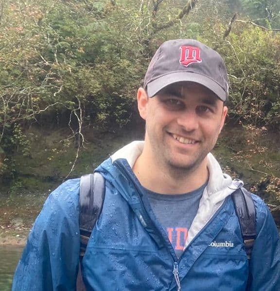 Smiling man in a blue rain jacket and cap stands outdoors in front of trees and a river.