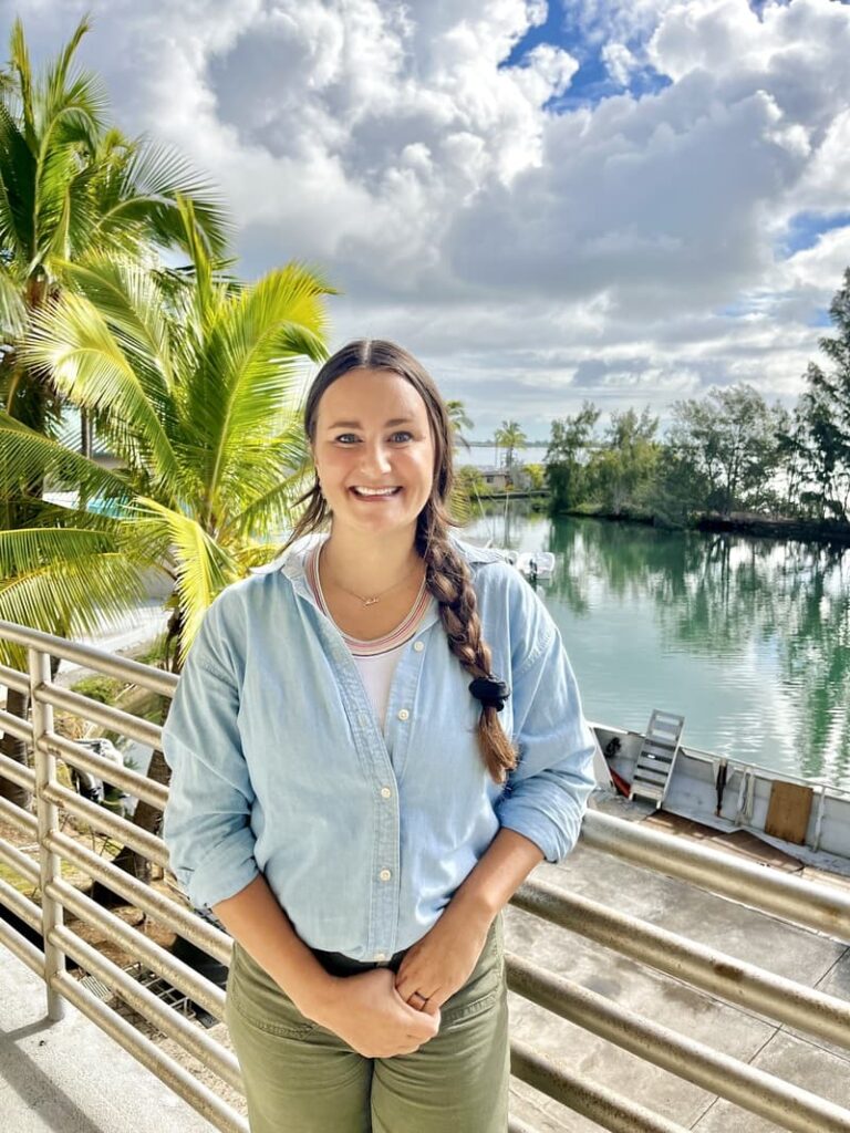 Smiling woman with a braid stands by a railing, palm trees and water in the background on a sunny day.