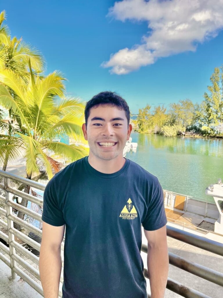 Smiling man in a black t-shirt stands by a waterfront with palm trees and boats under a blue sky.