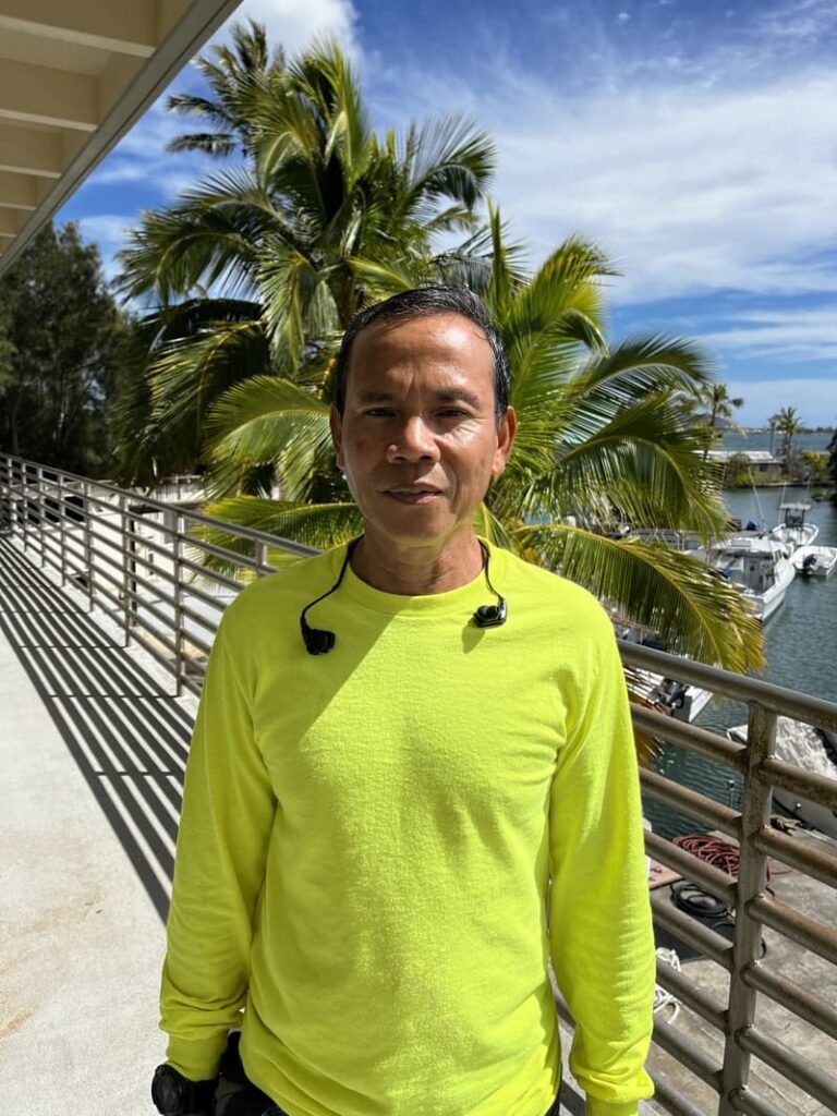 Man in a bright yellow shirt stands on a sunny balcony near palm trees and water, looking at the camera.