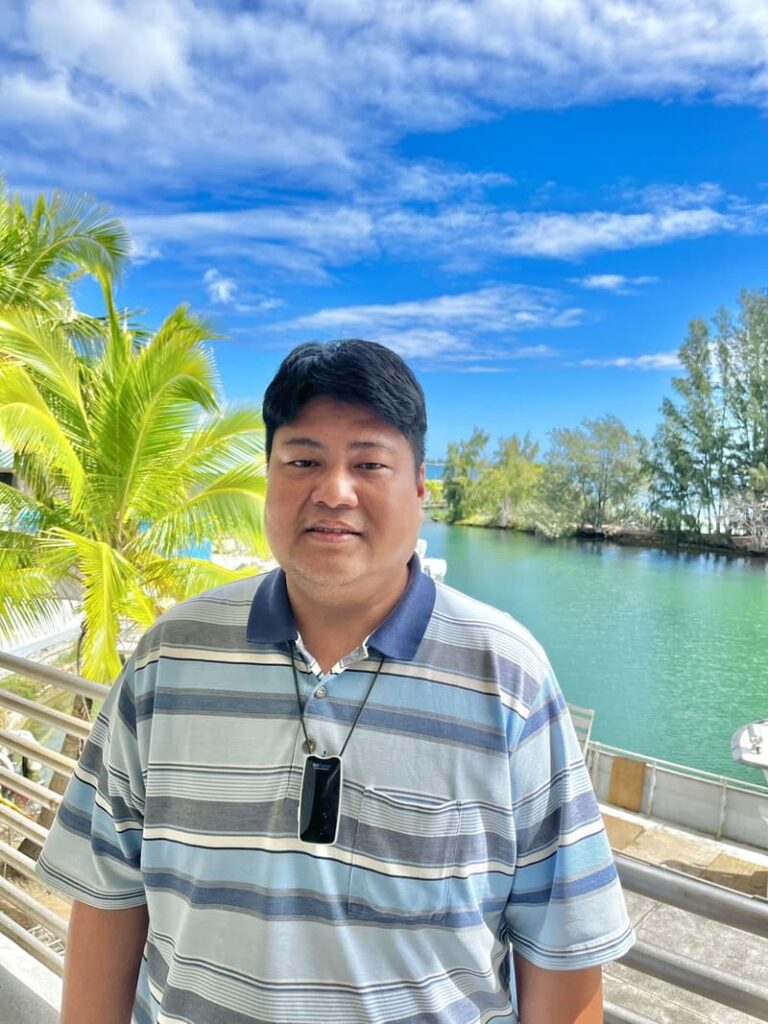 Man in a striped shirt stands by a waterfront with palm trees and a bright blue sky in the background.