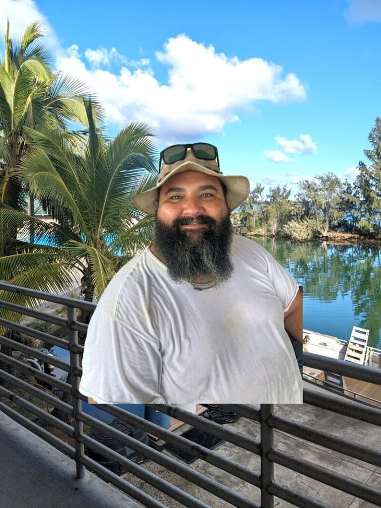 A smiling man in a hat and sunglasses stands by a railing near water, palm trees, and a blue sky.