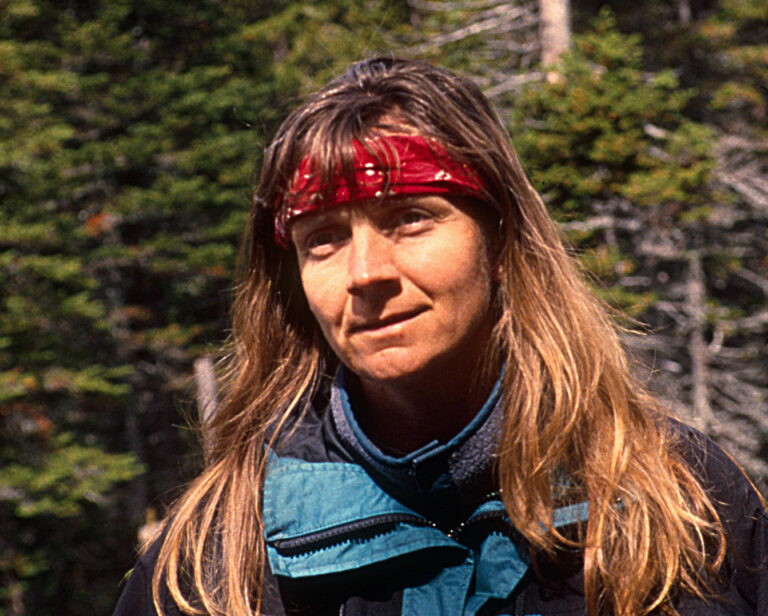 A woman with long hair and a red bandana stands outdoors, wearing a blue and black jacket, with trees behind her.